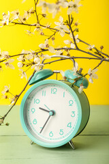 Blooming spring branches and blue alarm clock on green table near yellow wall