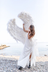 Beautiful angel with big white wings. Young tender woman in white silk dress walking at seashore