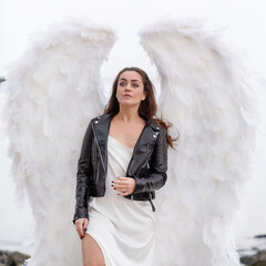 beautiful young woman with white wings. White angel in leather jacket and in white silk dress. holy woman 