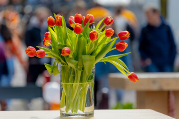 Colourful of King’s Day (in Dutch: Koningsdag) Selective focus a vase of orange tulips on the table along street with blurred peoples walking on street as background, National holiday in Netherlands.
