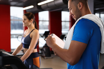 Fototapeta na wymiar Happy trainer writing down workout plan while woman doing exercise in modern gym