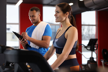 Fototapeta na wymiar Happy trainer writing down workout plan while woman doing exercise in modern gym, space for text