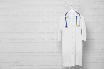 Medical uniform and stethoscope hanging on rack near white brick wall. Space for text