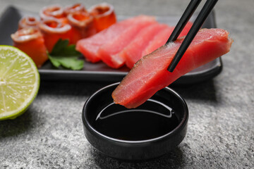 Chopsticks with raw tuna slice under soy sauce at grey table, closeup. Sashimi set served with lime