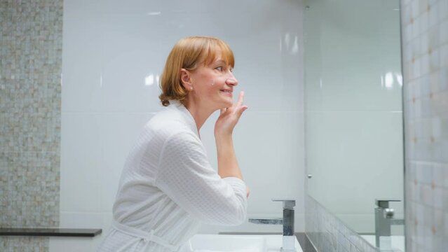 Caucasian senior woman looking at mirror then apply lotion on face.