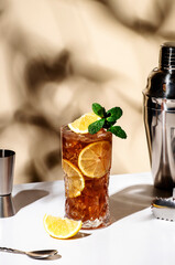 Long Island ice tea cocktail drink with vodka, rum, tequila, gin, liquor, lemon juice, cola and ice with lemon slice and mint in highball glass. Beige background, hard light, bar tools