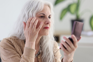 Mature woman with long grey hair examines wrinkles on face
