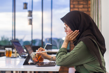 young woman in hijab sits working in a cafe using digital tablet and keyboard, next to her are drinks and healthy food