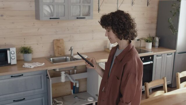 Mature Caucasian woman standing in kitchen searching for plumbing repair service phone number in Internet