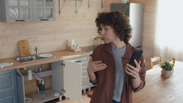 High angle medium shot of Caucasian woman standing in kitchen making video call to professional plumber