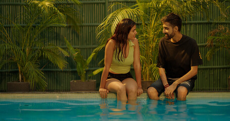 Happy young Indian marriage couple sitting edge of swimming pool water relax talk flirt together at resort Asian boyfriend girl friend smile discuss plans look each other enjoy fun joy weekend moment