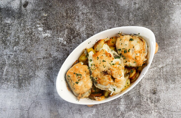 Baked cod with potatoes and herbs in a white baking dish on a dark grey background. Top view, flat lay