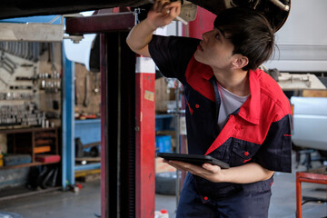 Professional young Asian male motor mechanic inspects undercarriage of electric car(EV) lifted by forklift jack for repair at garage, automotive maintenance service works industry occupation business.