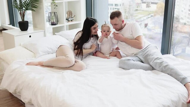 Happy affectionate family lying on king bed and hugging little cutie in white splendid room with city panorama view. Young father kissing sweet daughter in head while mother gently touching.