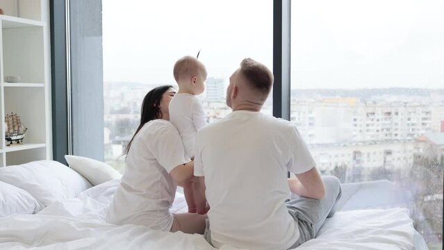 Rear view of loving father kissing his baby daughter while little girl hugging caring mom in morning after waking up. Young married couple with kid resting on white blanket in bedroom of modern flat.