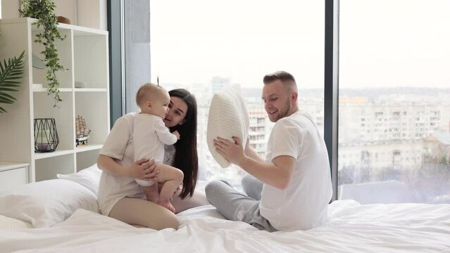 Relaxed young parents playing hide and seek using a pillow with baby daughter in bedroom interior. Charming mature adults and curious kid making most of leisure time at home.