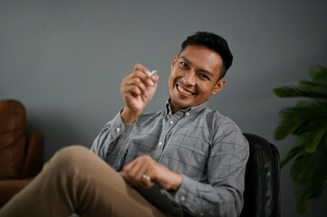 Cheerful Asian man sits on a black leather chair and points a pen at the camera.
