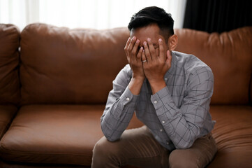 Stressed and depressed Asian man is covering his face, crying on the sofa in a living room