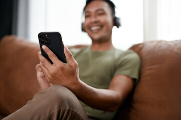 Happy Asian man using his smartphone and relaxing on a sofa in his living room.