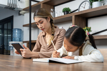 Beautiful mother teaching her lovely daughter to do homework in living room