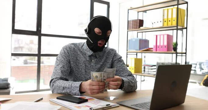 Excited man in robber mask talks into headset microphone sitting at table with computer in office. Emotional hacker counts money shrugging nervously