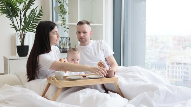 Sweet little baby drinking from sippy cup while joyful brunette woman snuggling with blonde male in cozy bedroom with tray on foreground. Caring family spending quality time together at home.