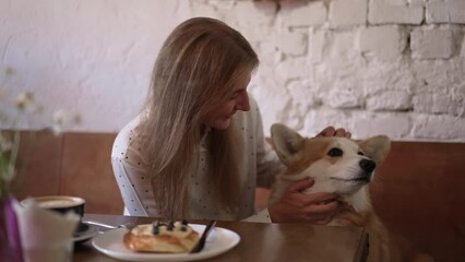 4k Woman strokes corgi dog and sits in cafe spbd. Portrait of young beautiful female touching cute pet and looking with happy smile, sitting at table with coffee in room. Red-haired lady owner touches