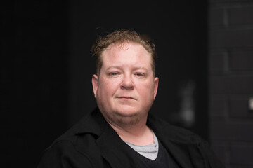 Fototapeta premium A disabled man celebrates his identity as a disabled and trans man, sitting in his powerchair with a black brick wall in the background, and expressing a direct, professional gaze.