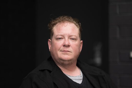 A Disabled Man Celebrates His Identity As A Disabled And Trans Man, Sitting In His Powerchair With A Black Brick Wall In The Background, And Expressing A Direct, Professional Gaze.