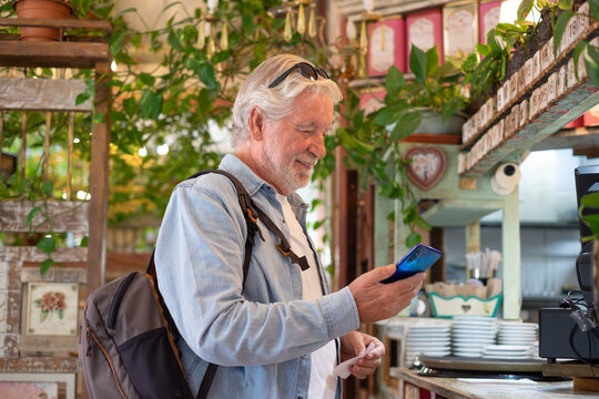 Smiling senior white-haired man pays at the coffee shop checkout using the application on his mobile phone. Concept of modernity and online electronic payments