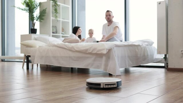 Brunette woman playing with infant girl on bed while young man turning on robot vacuum cleaner via remote control in studio room. Modern family resting at weekend using one touch home cleaning.