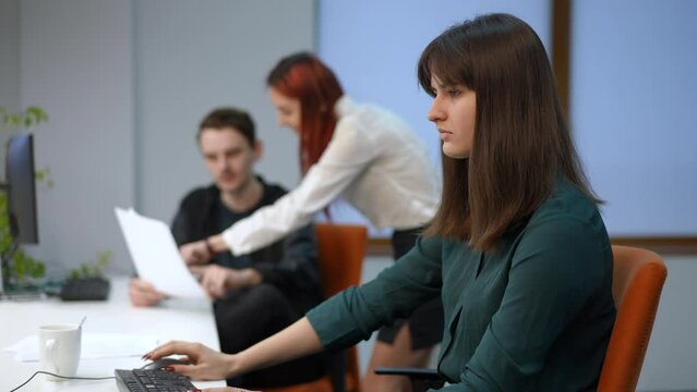 Side view concentrated young woman working in office with blurred colleagues talking at background. Intelligent beautiful Caucasian employee sitting at table with computer typing on keyboard