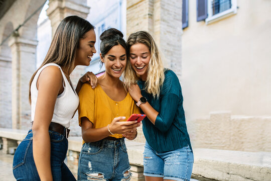 Three diverse young women having fun using mobile phone together in city street. Technology lifestyle and social media concept. Generative AI