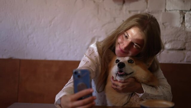 4k Woman takes selfie with dog and sits in cafe spbd. Close view of young female taking photo and kissing adorable pet, holding smartphone and sitting at table with coffee in interior. Beautiful lady
