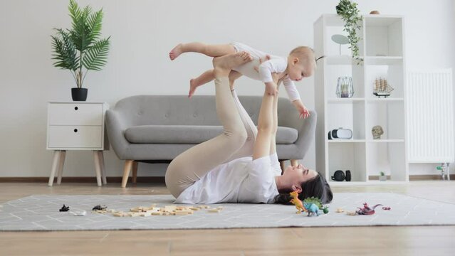 Beautiful brunette woman holding infant girl high in air with arms and legs while lying on carpet in living room. Loving mother letting small daughter fly safely during their funny games.