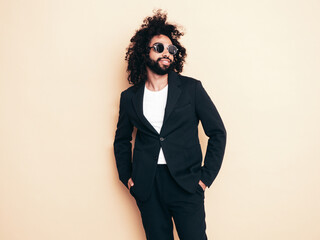 Handsome smiling hipster model.  Unshaven Arabian man dressed in summer suit jacket clothes. Fashion male with long curly hairstyle isolated on beige in studio. Cheerful and happy. In sunglasses