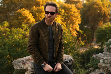 A bearded Caucasian man with sunglasses and a dark green jacket posing in front of a blurry autumn background	