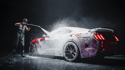Naklejka premium Automotive Detailer Washing Away Smart Soap and Foam with a Water High Pressure Washer. Red Performance Car Getting Care and Treatment at a Professional Vehicle Detailing Shop