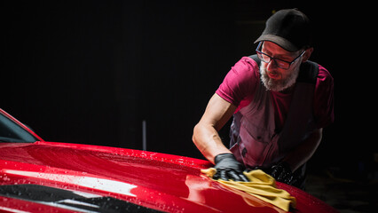 Car Wash Expert Using a Microfiber Cloth to Dry Up a Red Modern Sportscar. Adult Man Cleaning Away...