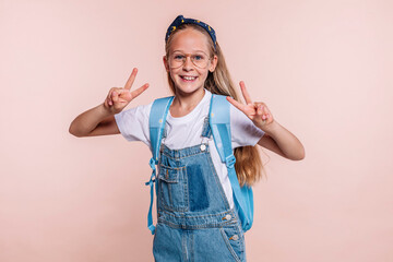Blond school girl with blue eyes, wears a blue backpack, eyeglasses and denim overalls, does peace...