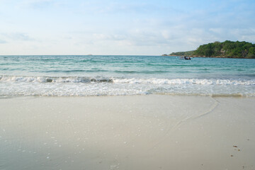 Beach at sea view with waves splashing or crash the sand with sunlight on sunny summer holiday with floating pier, island and blue sky background.