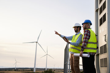 Team two young maintenance engineers working on wind turbine at wind farm. Male technician using tablet and pointing out one of windmills to professional colleague. Copy space - Powered by Adobe