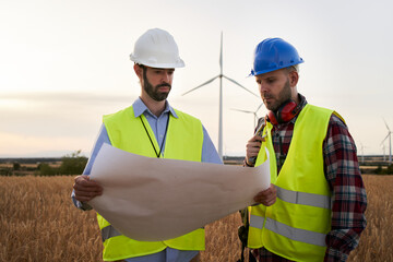 Two wind engineers wearing vests and helmets look at plans of wind turbine system in field. Professional technicians outside a wind power plant. Clean renewable energy concept