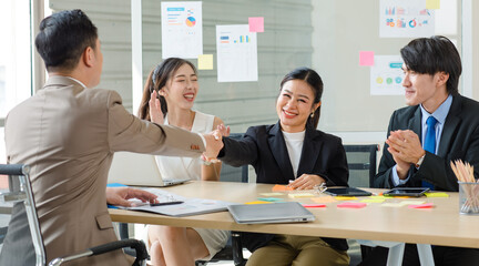 Asian professional successful businesswoman sitting smiling shaking hands greeting with unrecognizable businessman in formal business suit while male female colleagues clapping hands celebrating