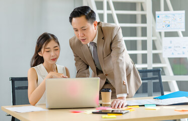 Asian professional successful male businessman manager mentor in formal business suit standing helping teaching supporting female businesswoman colleague sitting working with laptop notebook computer