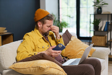 Tired man in feeling headache while holding hand on head with closed eyes. He sit home with mobile...
