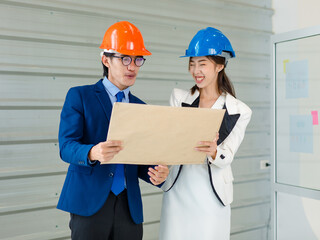 Asian professional successful female businesswoman and male businessman engineering employees in formal business suit and safety helmet standing holding blueprint and water level gauge discussing