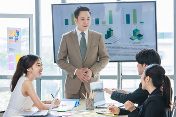 Asian professional successful male businessman lecturer speaker presenter in formal business suit standing at strategy data information monitor speaking to audiences in company office meeting room