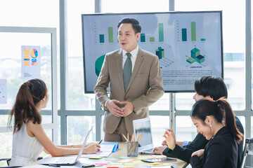 Asian professional successful male businessman lecturer speaker presenter in formal business suit standing at strategy data information monitor speaking to audiences in company office meeting room