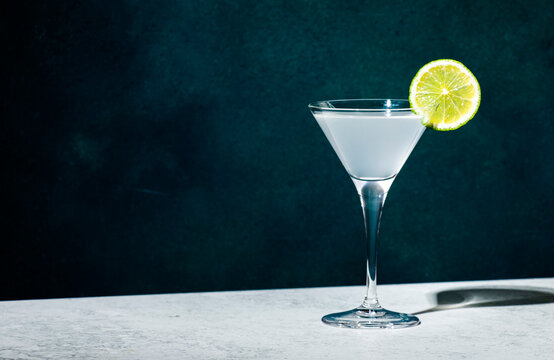 Vodka gimlet alcoholic cocktail with vodka, sugar syrup, lime juice and ice in martini glass, dark green background, hard light and shadow pattern, minimalism